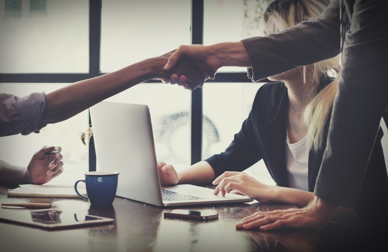 345349079 A seated person is shaking hands with a standing person while another person types on a laptop. The standing person has one hand on a desk, which also has a coffee cup
